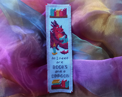 Photograph of cross stitched bookmark, showing a dragon reading a book, and the words 'All I need are BOOKS and a DRAGON'. Shown on rainbow fabric background.